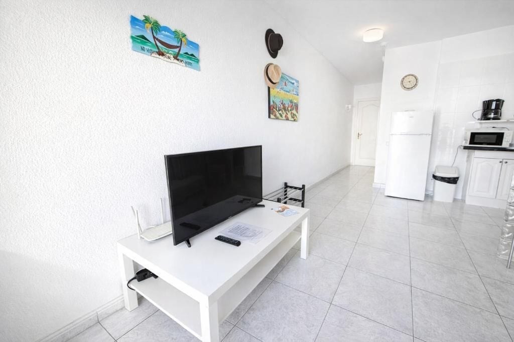 Un salón moderno con un televisor de pantalla plana sobre una mesa blanca, un mueble blanco pequeño y una zona de cocina con microondas y refrigerador.