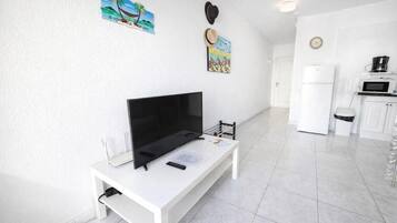 Un salón moderno con un televisor de pantalla plana sobre una mesa blanca, un mueble blanco pequeño y una zona de cocina con microondas y refrigerador.