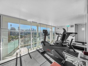A gym with cardio equipment and a city view through large windows. - Harbour Muse - Stylish 2 or 3 Bedroom Apartment with Spectacular Harbour Views (Darlinghurst)