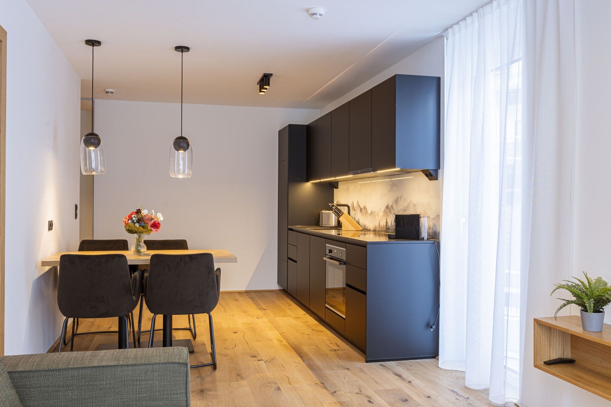 A modern kitchen with dark cabinetry, a dining area with a wooden table and chairs, pendant lighting, and a view of a mountain landscape through the window.