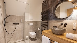Et moderne badeværelse med et glasafskærmet brusebad, toilet, en trævask med skålvask i sten og et rundt spejl.