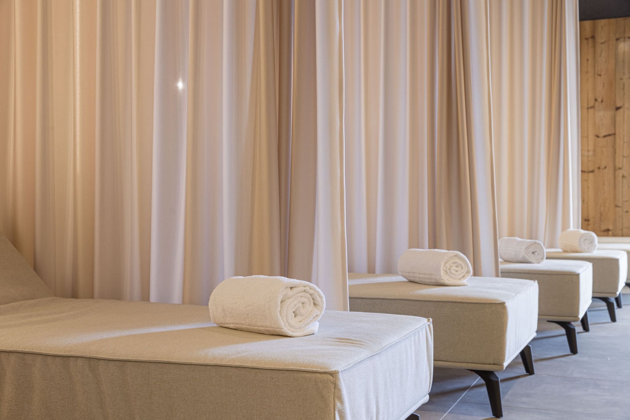 Eine Reihe von Wellnessliegen mit Handtüchern, sandfarbenen Vorhängen und einer Holzwand.