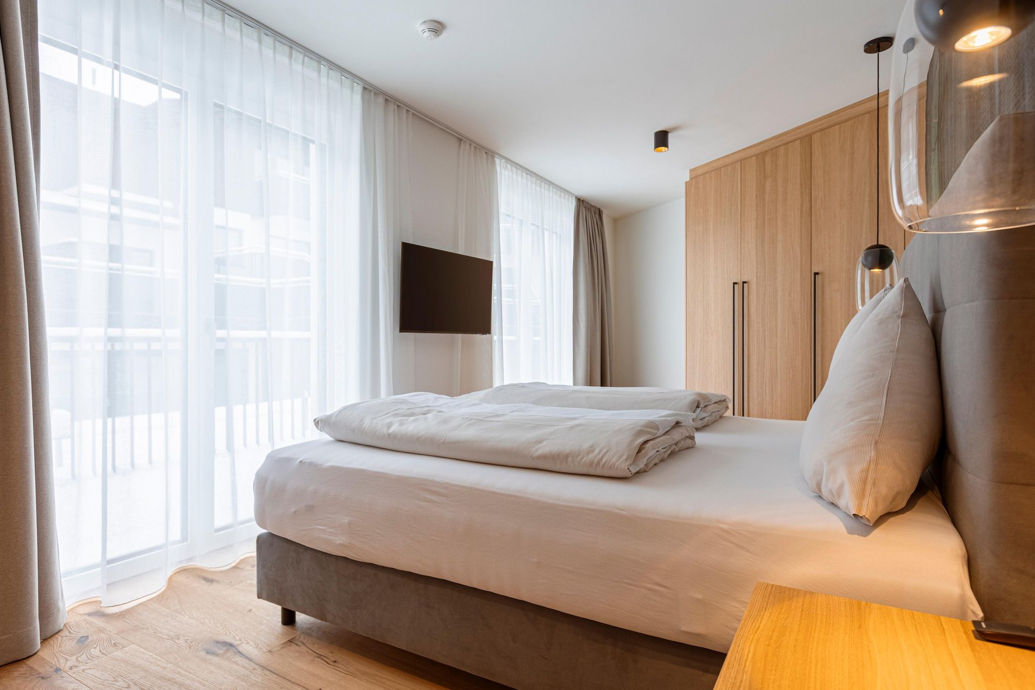 A modern hotel room with a large bed, a flat-screen TV mounted on the wall, and a wooden wardrobe.