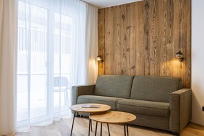 A room with a wooden wall, a grey sofa, two round wooden tables, and a window with sheer curtains. - Fiss – Holiday apartment in the Alps (Fiss)