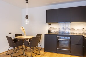 A modern kitchen with dark cabinetry, a central island with an oven, and a dining area with a round table and chairs. - Fiss – Holiday apartment in the Alps (Fiss)