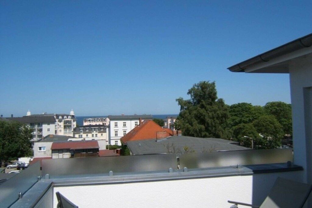 A residential building with a flat roof, a balcony, and a view of other buildings and trees.