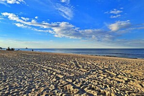 En sandstrand med fodspor, der fører hen til vandet, en klar himmel og enkelte mennesker i det fjerne.