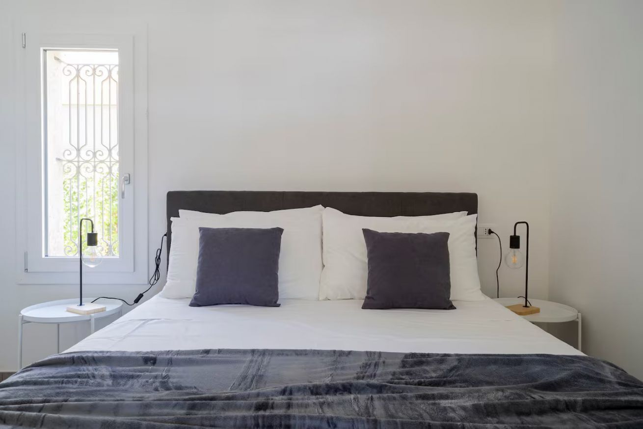 Un letto ben rifatto con una coperta a motivi, due comodini con lampade e una finestra con una griglia decorativa in metallo.