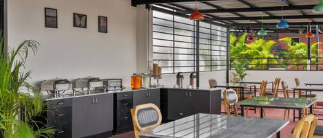 A modern cafe with a kitchen area, dining tables, chairs, and a view of greenery through large windows.