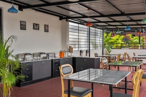 A modern cafe with a kitchen area, dining tables, chairs, and a view of greenery through large windows. - Wandr Hotel HSR Layout Sector 7 (Bengaluru)