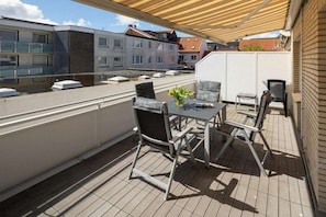 Ein überdachter Außenbereich mit Tisch und Stühlen, einem Blumentopf und Blick auf die Nachbargebäude.
