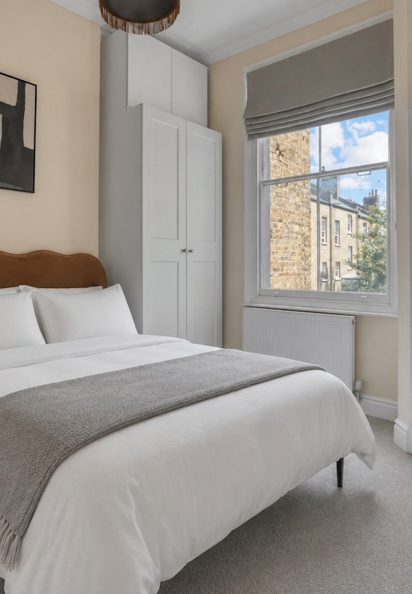 A bedroom with a bed, a wardrobe, a window with a view of buildings, and a radiator. - Stay Soho | Stylish 2BR Steps from the West End (London)