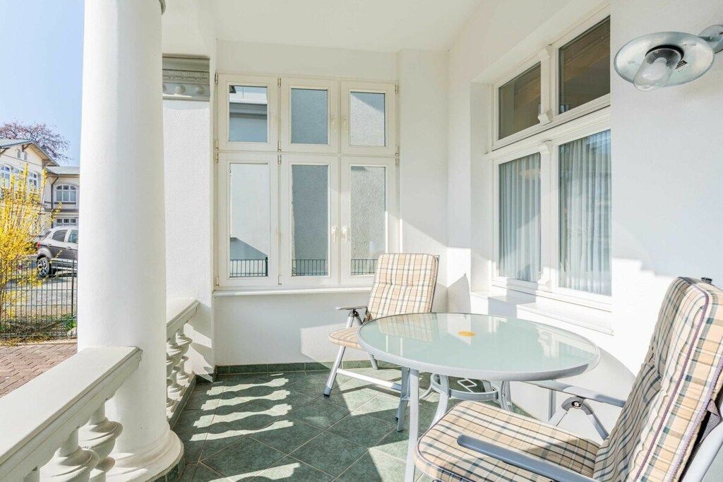 Een zonnig balkon met een ronde glazen tafel en twee gestreepte stoelen, een witte kolom en een raam met veel ruitjes.