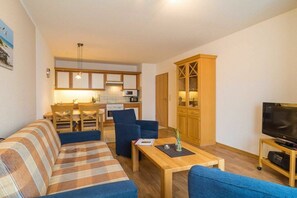 A living room with a sofa, a coffee table, and a television. There is a kitchen area in the background with cabinets and a dining table. - House by the spa park (Ahlbeck (Seebad))