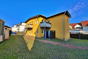 Un edificio amarillo con balcón, un patio empedrado y un coche azul estacionado al fondo.