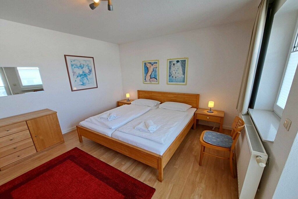 Een slaapkamer met een houten bed, een rood tapijt, een houten dressoir en twee kunstwerken in een lijst aan de muur.