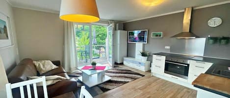 En moderne stue med sofa, sofabord og spisebord. Der er et køkken med komfur, ovn og skabe. Et fjernsyn er monteret på væggen, og et vindue giver udsigt til det fri.