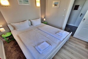 Ein Doppelbett mit weißer Bettwäsche, zwei Nachttische mit Lampen und eine Tür rechts.