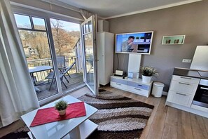 A modern living room with a sliding glass door to a balcony, a flat-screen TV, a dining table with a red placemat, and a kitchen area with cabinets and a countertop. - Holiday apartment with 1 bedroom (Heringsdorf (Seebad))