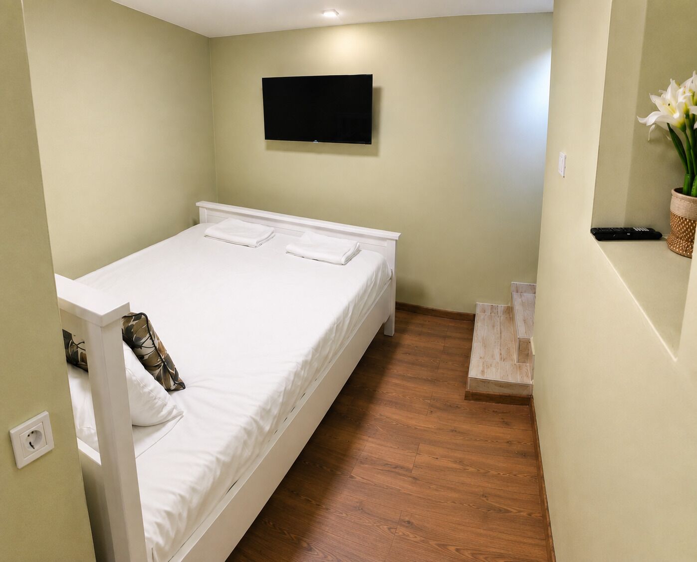 Ein Einzelbett mit weißer Bettwäsche, ein an der Wand befestigtes Fernsehgerät und ein Holzboden.
