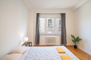 Ein Schlafzimmer mit einem Bett, einem Heizkörper, einem Fenster mit Vorhängen, einem Nachttisch mit Lampe und einer Topfpflanze.