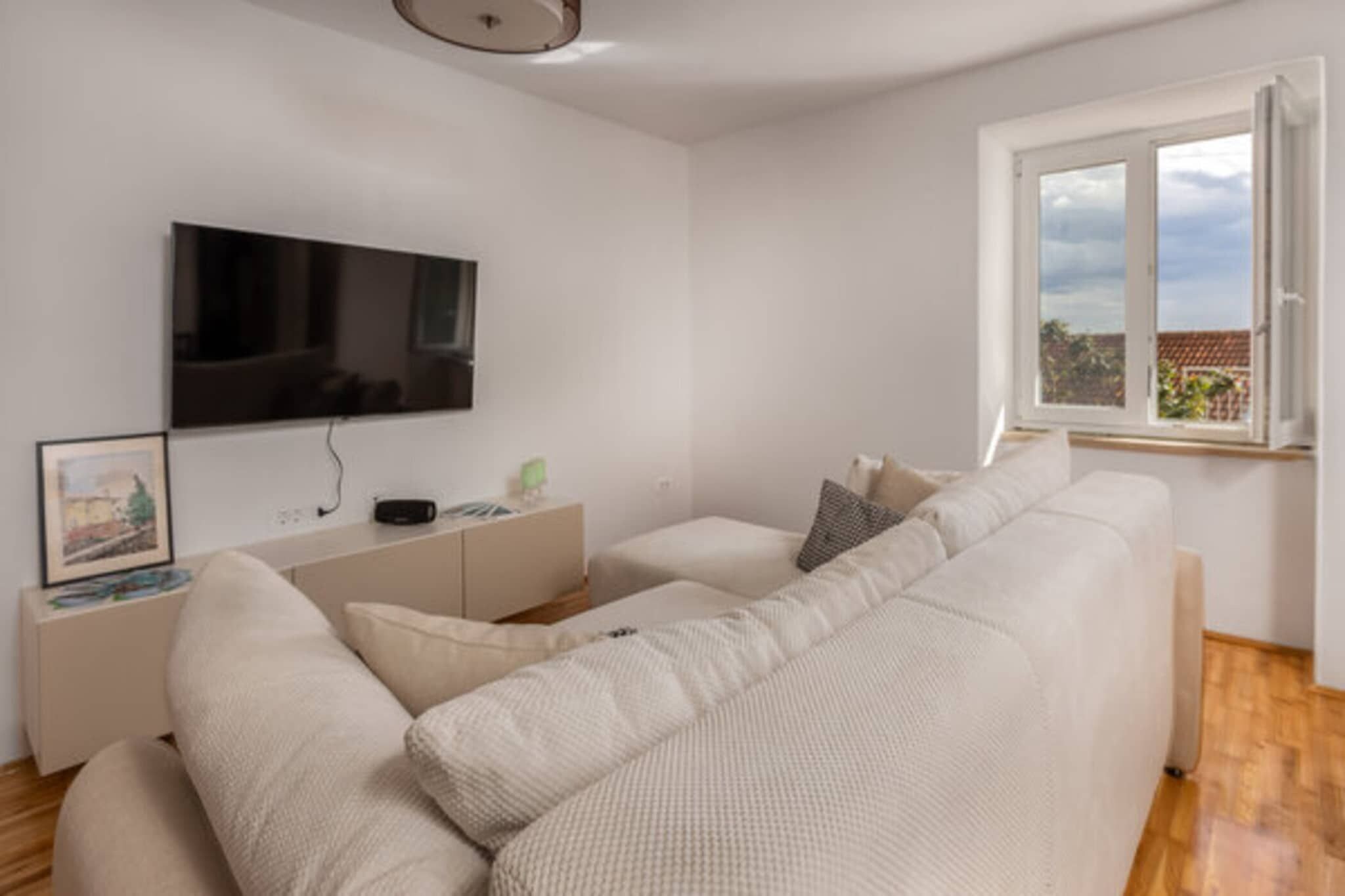 A modern living room with a large flat-screen TV mounted on the wall, a white sofa with cushions, a small side table, and a window with a view of the outside.