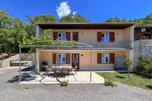 Ein zweigeschossiges Gebäude mit überdachter Terrasse, Sitzgelegenheiten im Freien und einem kleinen Garten.