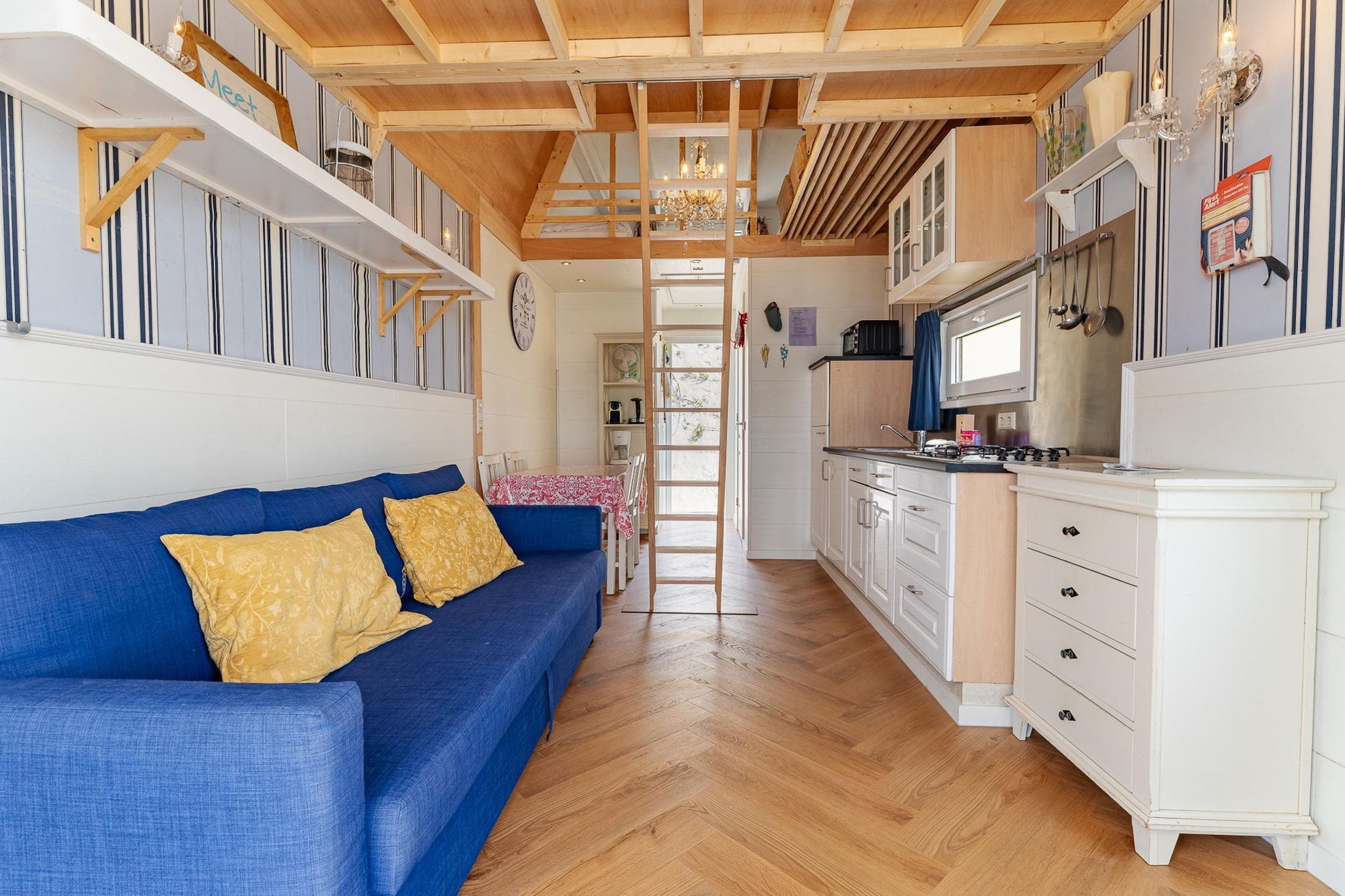 Een knus woongedeelte met een blauwe bank, houten vloeren, een keuken met witte kasten en een houten plafondconstructie.