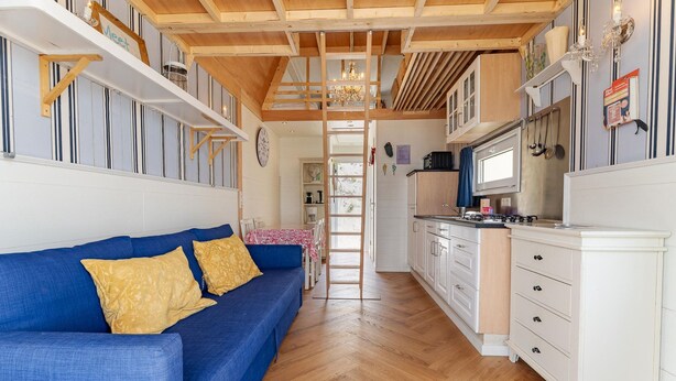 Ein kompakter Wohnraum mit blauem Sofa, Holzboden, Küchenzeile mit weißen Schränken und einer Holzbalkendecke.