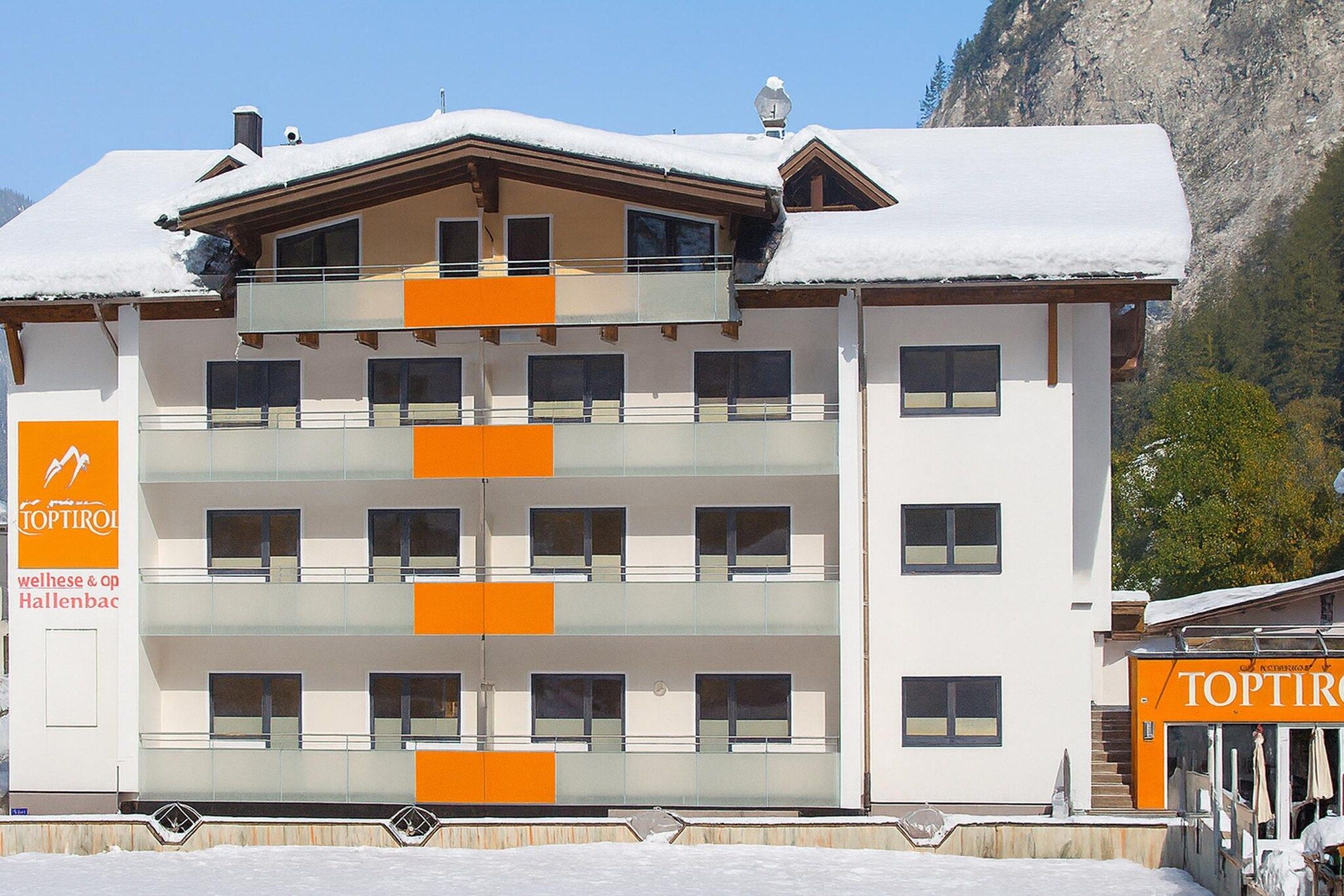 A multi-story hotel with balconies, a sign for 'Topirol' on the left, and a mountain backdrop.