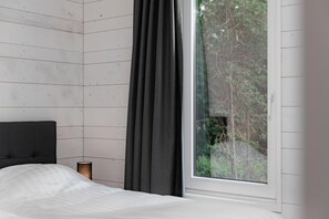 Een slaapkamer met een bed, nachtkastje, lamp en raam met uitzicht op bomen.