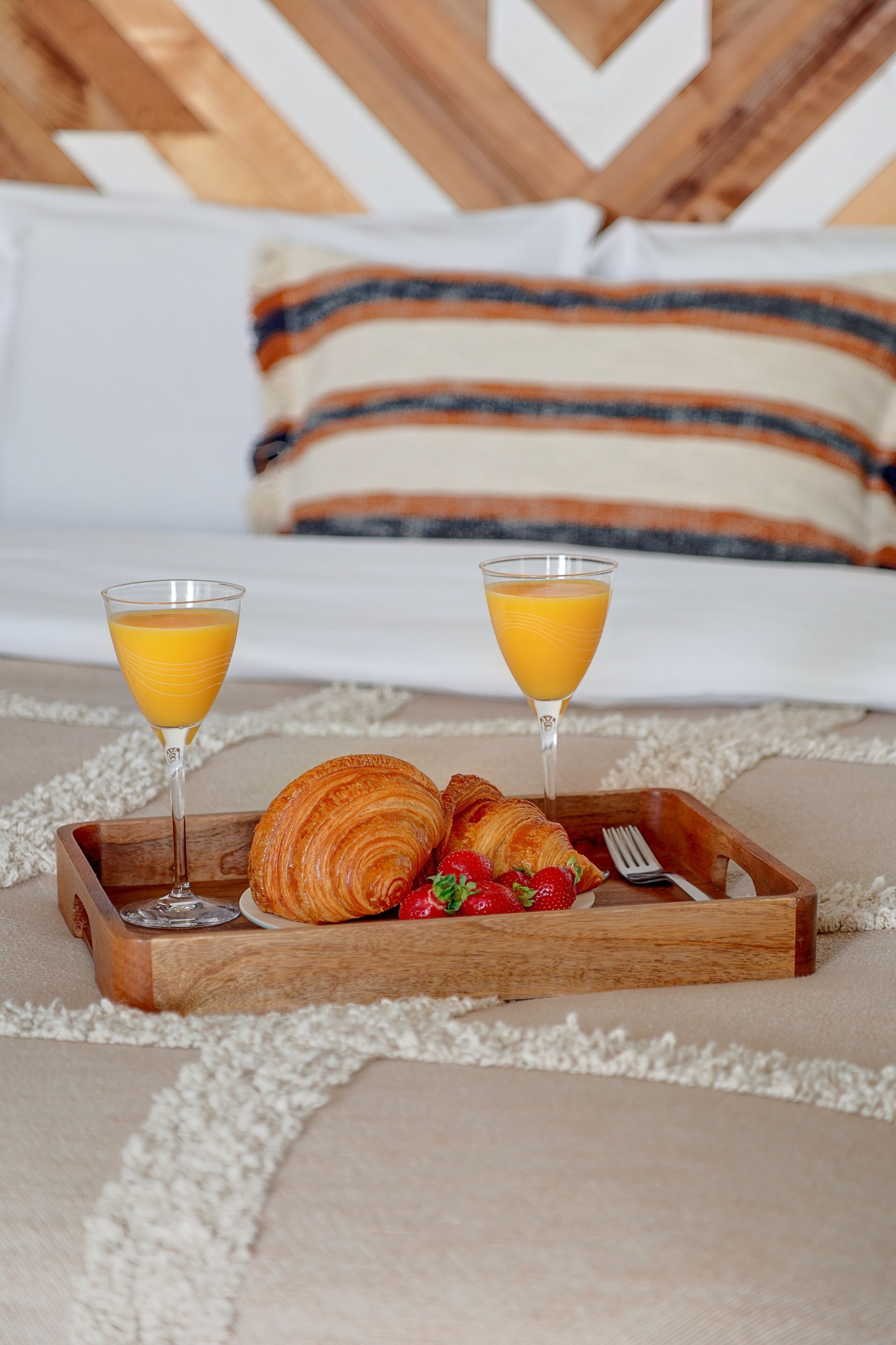 Een ontbijfwagen met croissants en aardbeien, twee glazen sinaasappelsap, en een vork op een bed met een gestreept kussen.