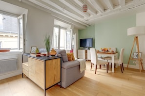 A modern living room with a grey sofa, a wooden cabinet, a dining area with a table and chairs, a television, and a window with a view of the outside. - Charming Stay Near Louvre & Palais Royal (Paris)