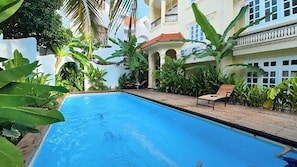 A swimming pool with a tiled deck, surrounded by lush greenery and a multi-story building with balconies.