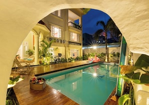 Una piscina interior con cubierta de madera, rodeada de plantas y un edificio con balcones.