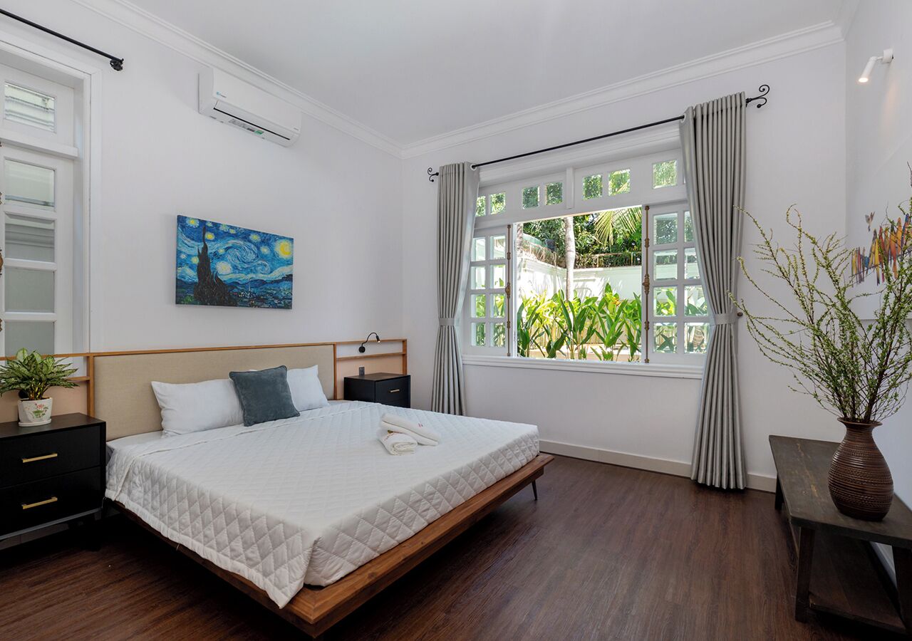 Une chambre à coucher avec un lit, des tables de chevet, un tableau, une fenêtre avec des rideaux et une vue sur la verdure à l’extérieur.