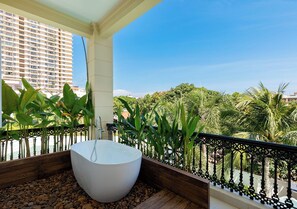 A modern bathroom with a freestanding bathtub, surrounded by a wooden deck and a metal railing, overlooking a lush green landscape and a high-rise building.