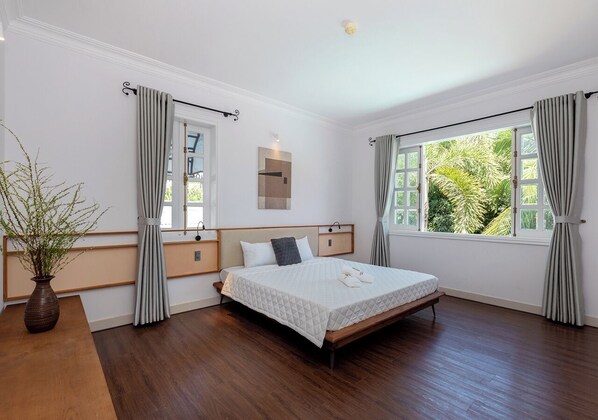 A bedroom with a bed, bedside tables, a nightstand, a window with curtains, and a plant.