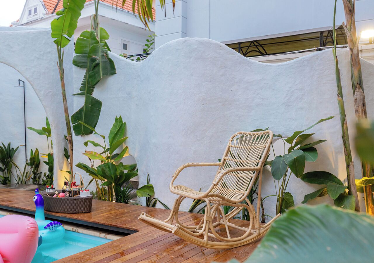 Un fauteuil à bascule en osier placé près d’une piscine, avec un mur blanc et des plantes tropicales en arrière-plan.