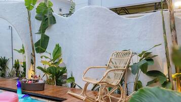 Un fauteuil à bascule en osier placé près d’une piscine, avec un mur blanc et des plantes tropicales en arrière-plan.