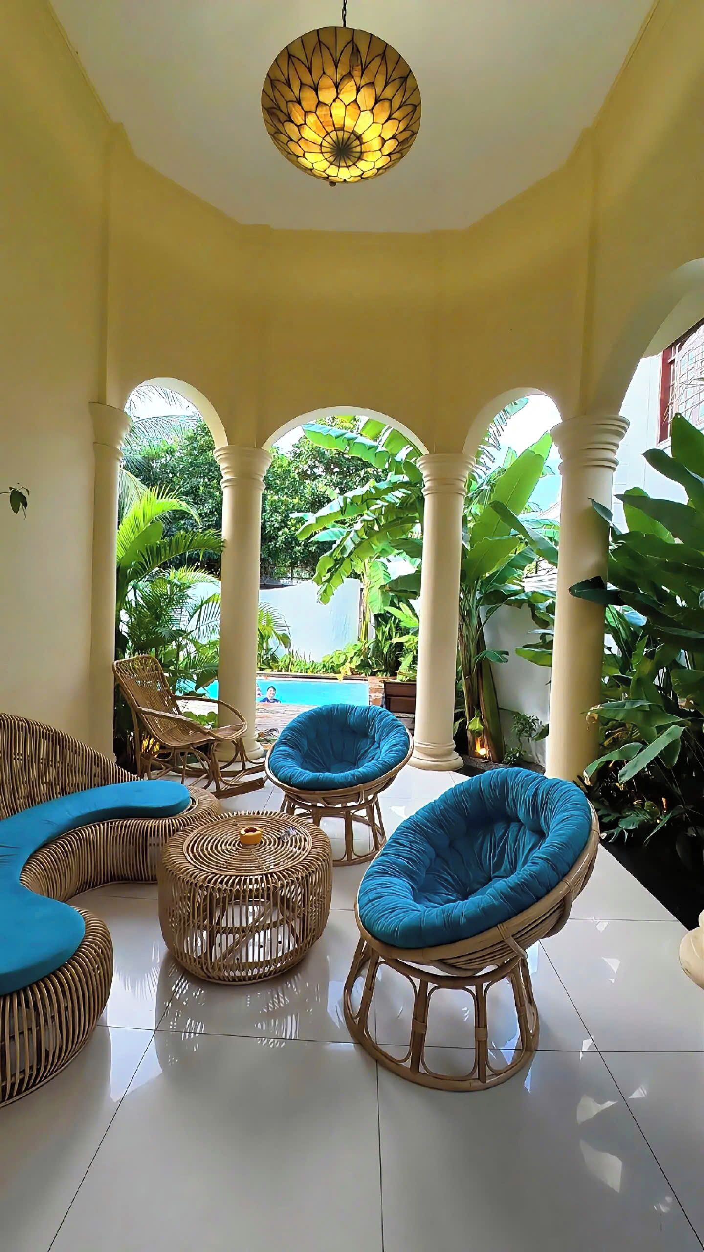 A sunlit indoor space with a pool visible through an archway, wicker furniture, and a hanging light fixture.