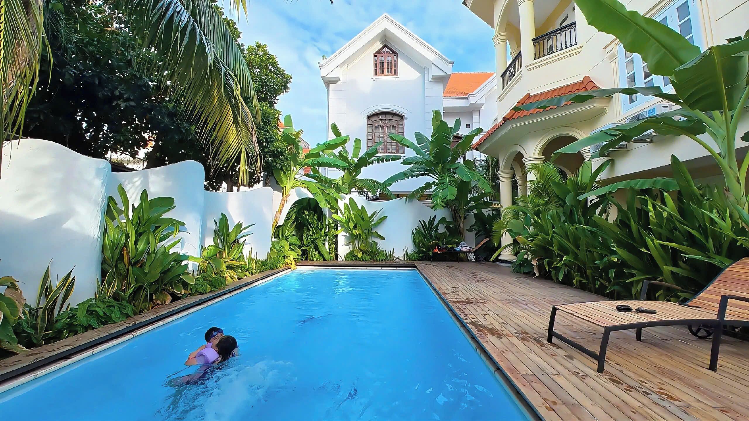 A swimming pool with a person swimming, surrounded by lush greenery and a building with a white facade and red roof in the background.