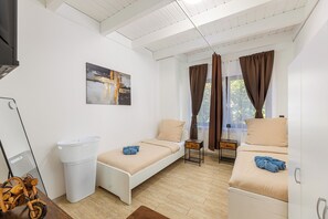 Ein Zimmer mit zwei Betten, einer weißen Wand, einem Gemälde, einem Fenster mit Vorhängen, einem Holzboden und einer weißen Mülltonne.