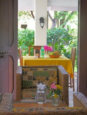 Un coin repas avec une table dressée pour deux personnes, une chaise en osier, une nappe jaune et une vue sur la verdure à l’extérieur.
