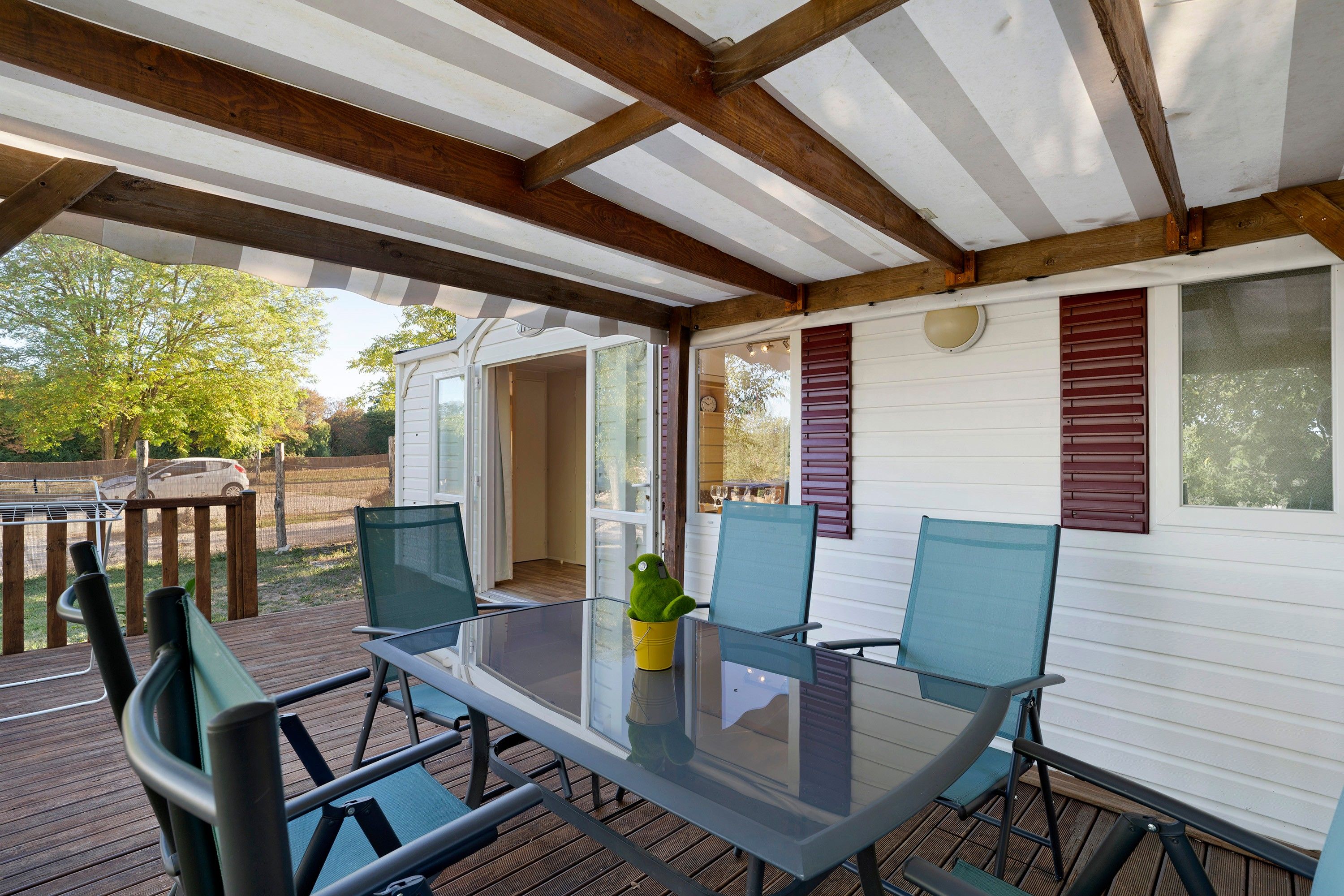 Une terrasse couverte avec une table et des chaises, une petite plante en pot et une vue sur une maison avec des volets.