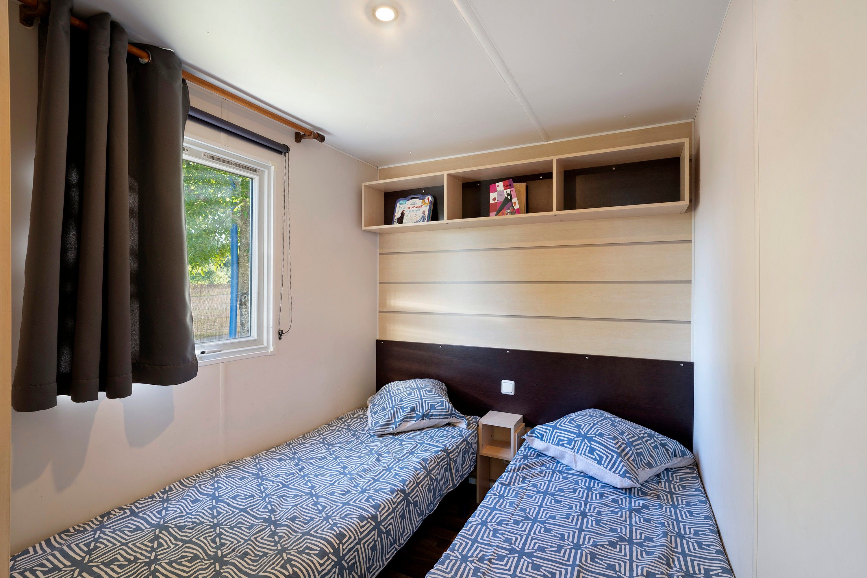 A compact bedroom with two single beds, a window with curtains, and built-in shelves above the beds.