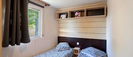 Un dormitorio compacto con dos camas individuales, una ventana con cortinas y estanterías empotradas sobre las camas.