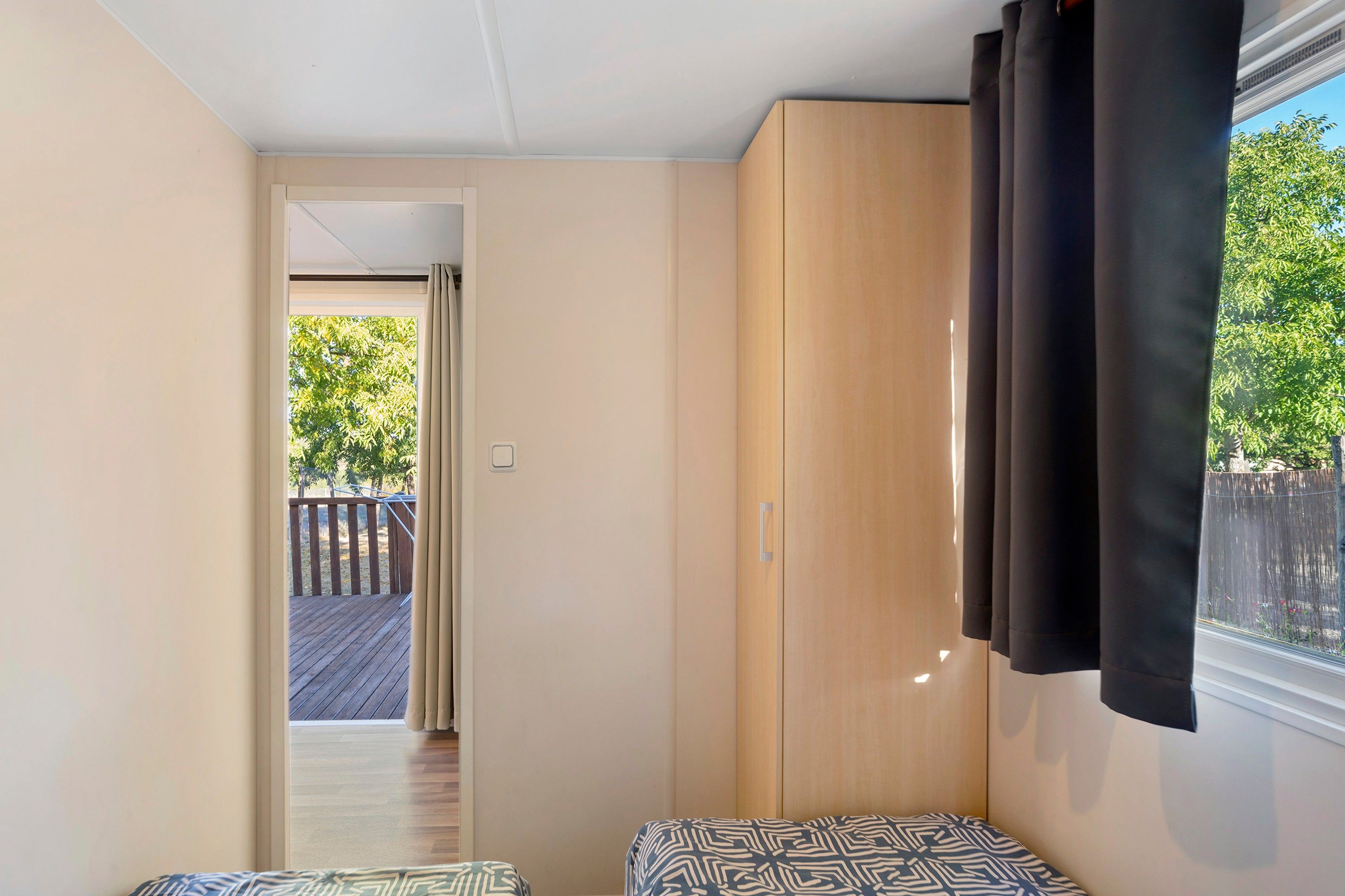 Un dormitorio con un armario de madera, una ventana con cortinas y vistas a una terraza y árboles.