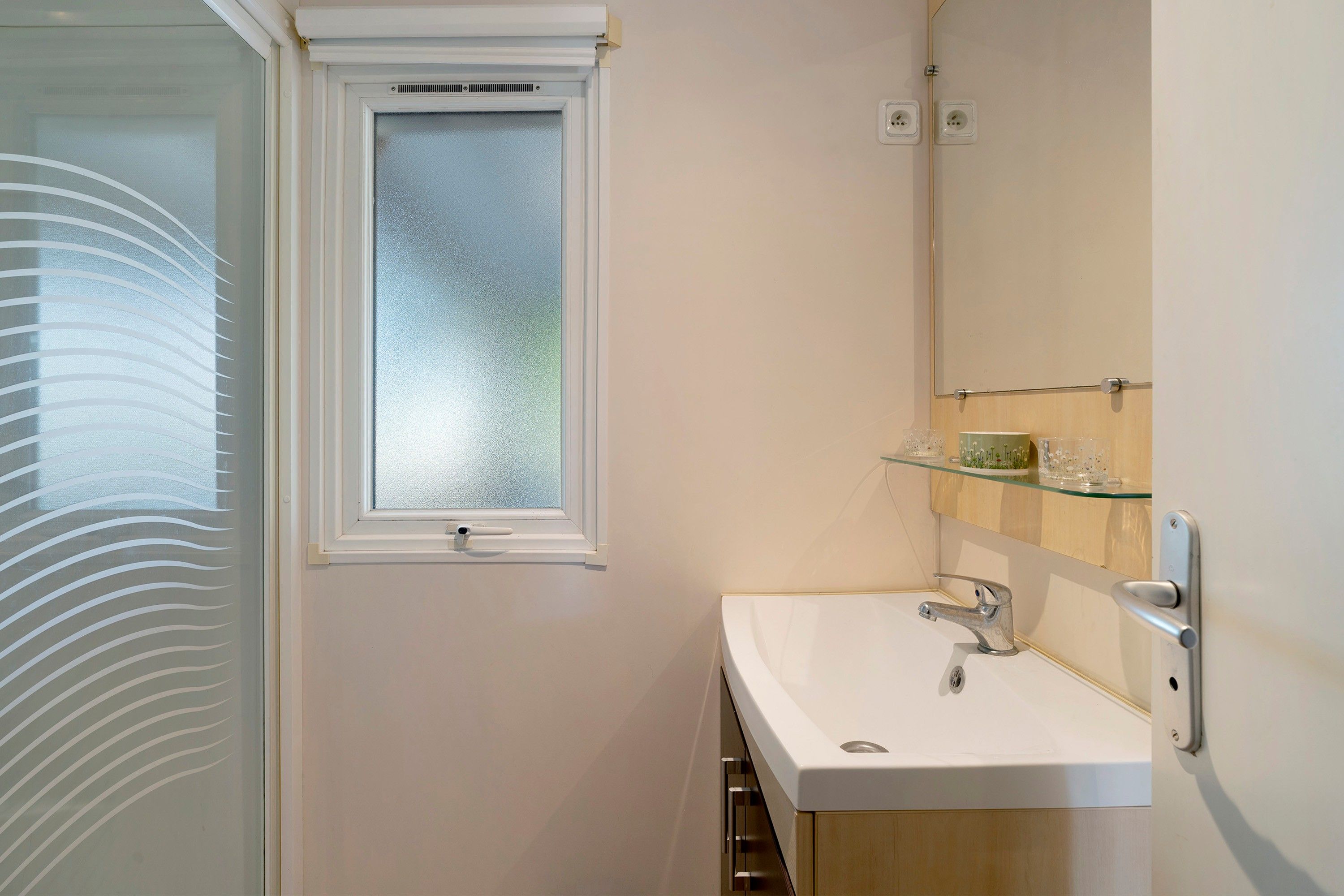 Un baño con un lavamanos blanco, un estante de vidrio encima y una ventana esmerilada, además de una puerta con un panel de vidrio satinado.