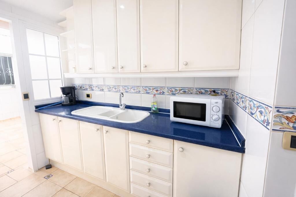 Una cocina con armarios blancos, encimera azul, pileta, microondas y ventana.